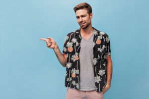 Stylish bright man in black shirt with print and plaid t-shirt showing his finger to place for text on blue isolated background..
