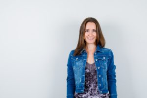 young lady posing while standing in blouse, denim jacket and looking cheery. front view.